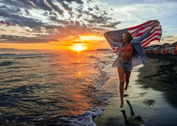 Happy woman holding american USA flag and running on the beach. Independence Day, travel concept